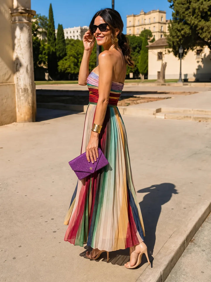 Side view of woman in a vibrant, pleated maxi dress, holding a purple clutch, in a sunlit outdoor setting.