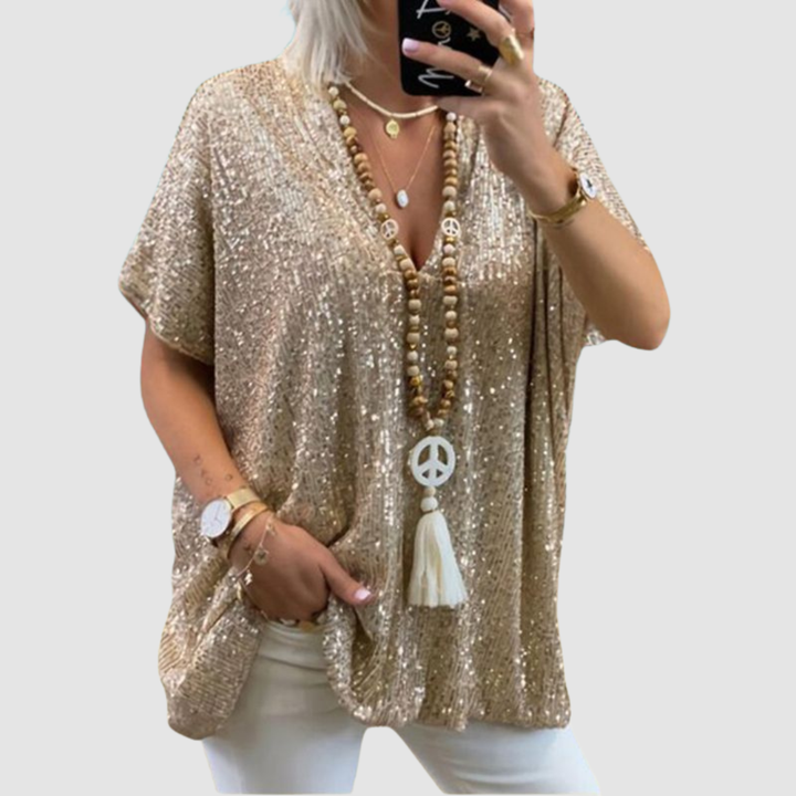 Woman wearing a gold V-neck sequin top with white pants, accessorized with layered necklaces and bracelets, on a neutral background.