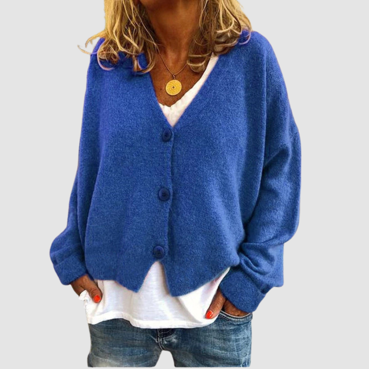 Woman wearing a blue knit cardigan over a white shirt and blue jeans, set against a plain gray background.