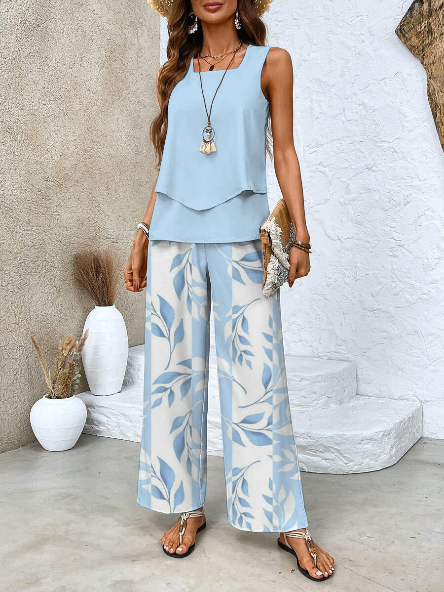 Woman in light blue two-piece set with layered top and leaf pattern pants, holding a straw clutch, standing outdoors near white steps.