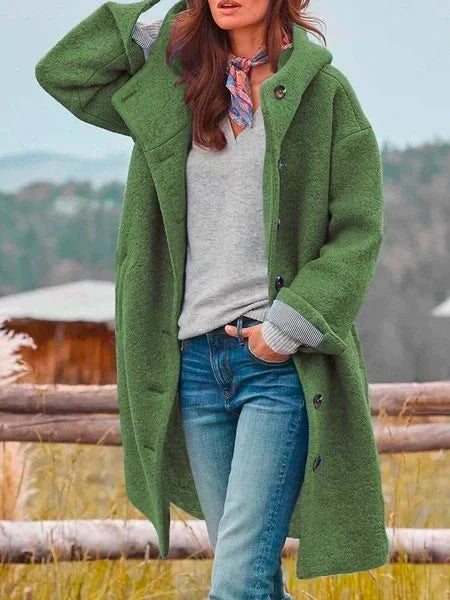 Woman in long green coat with buttons, posing near a wooden fence outdoors with scenic mountain backdrop.