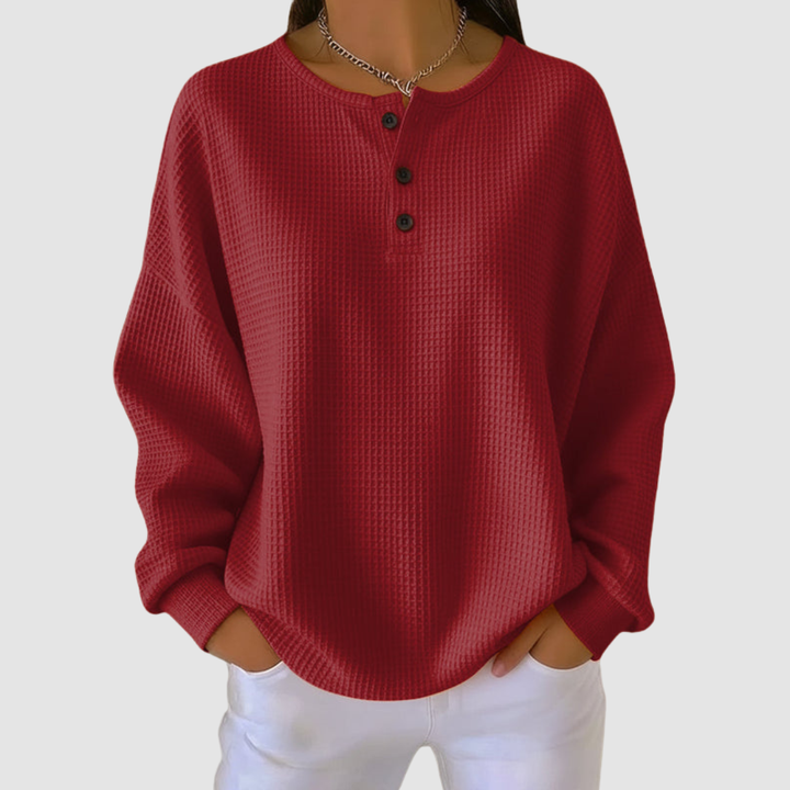 Red relaxed sweater with buttoned neckline and textured fabric, paired with white pants in a studio setting.