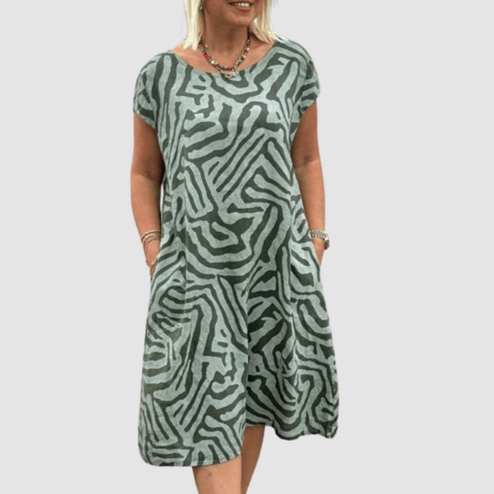 Woman wearing a green, vintage-inspired dress with abstract pattern, hands in pockets, against a plain backdrop.