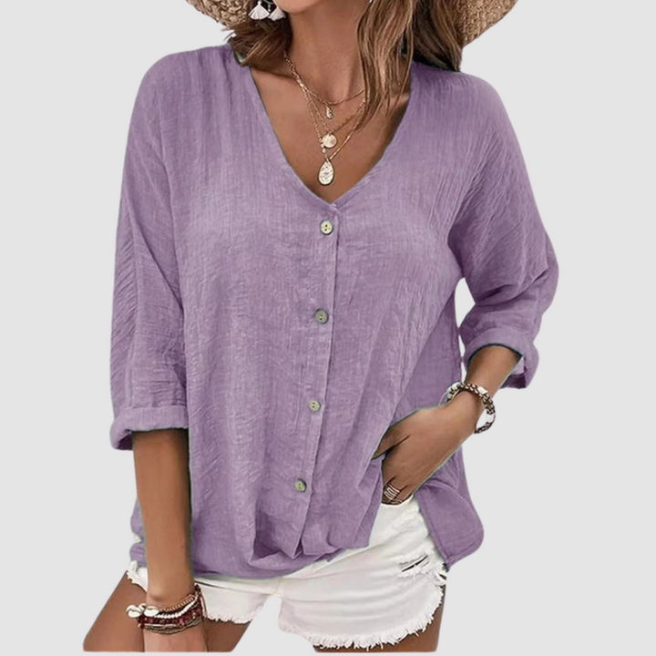 Woman wearing a purple V-neck blouse with buttons, accessorized with necklaces and bracelets, paired with white shorts against a plain background.