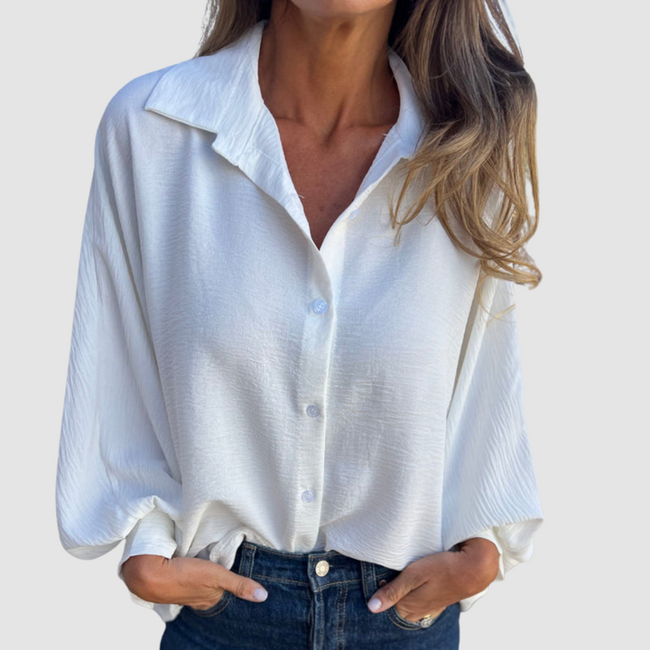 Model wearing a white oversized blouse with buttons, blue jeans, hands in pockets, gray background.