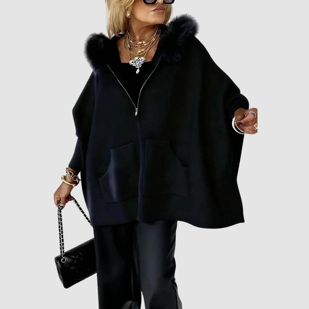 Woman wearing a black casual coat with a fluffy hood, large pockets, and black pants, holding a black handbag in a studio setting.