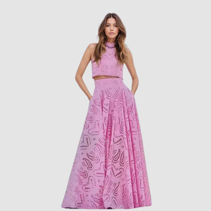 Woman in a pink two-piece set with intricate cutout design, posing with hands in pockets against a white background.