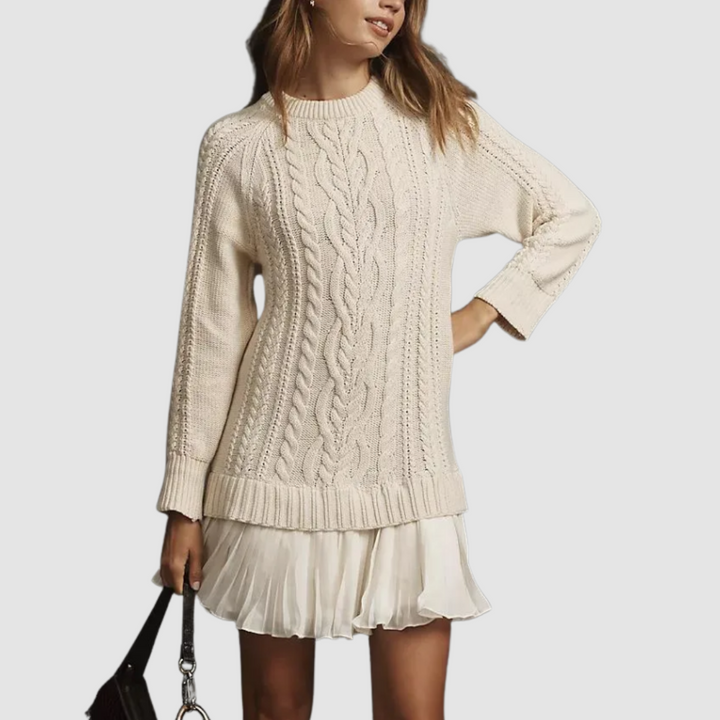 Cream cable knit mini dress with ribbed cuffs and a pleated hem, styled with a black handbag. Studio backdrop.