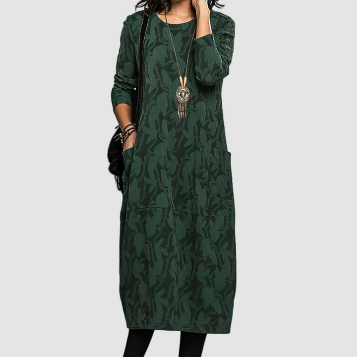 Woman wearing an elegant green dress with a botanical pattern, long sleeves, and a decorative necklace, set against a plain background.