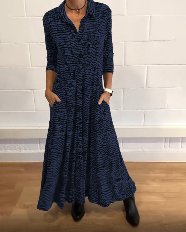 Navy-patterned maxi dress with 3/4 sleeves and collar, worn indoors against a white brick wall.