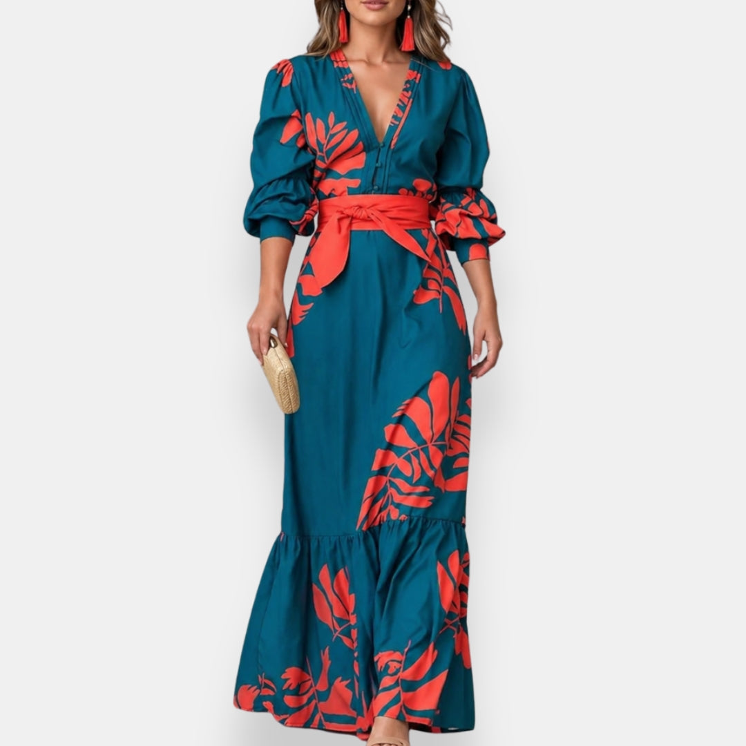 Woman wearing a teal maxi dress with bold red floral patterns and voluminous sleeves, holding a beige clutch in a studio setting.