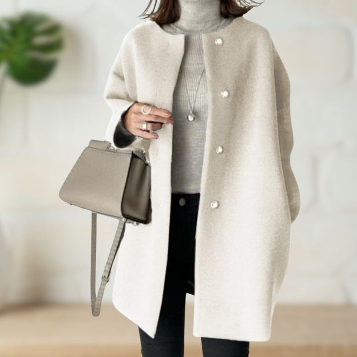 Cream mid-length coat paired with a light turtleneck and black pants, complemented by a beige handbag in a chic indoor setting.