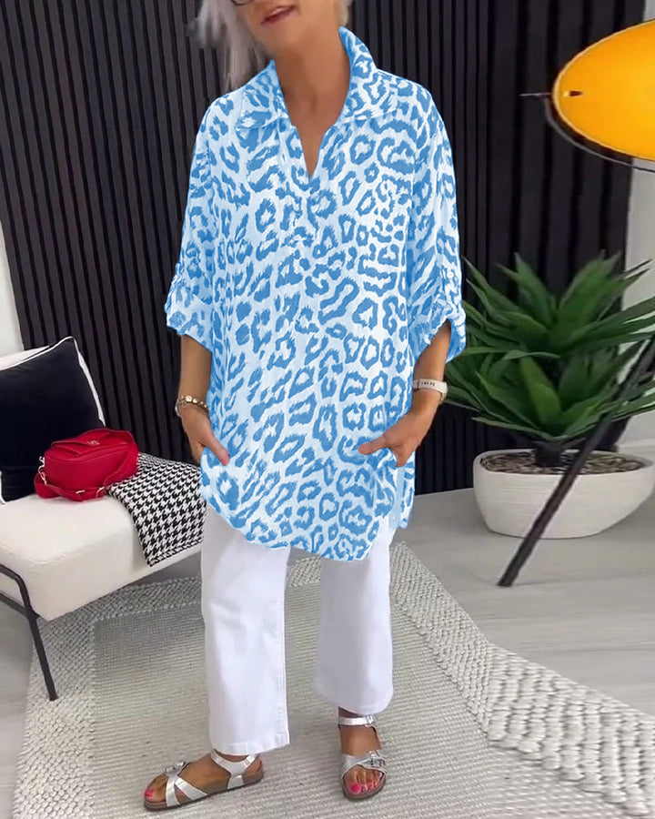 Blue leopard print blouse with collar, styled with white pants, in an elegant indoor setting featuring plants and decor.