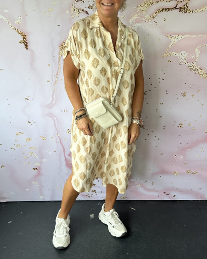 Woman in beige loose lapel collar dress with golden leaf prints, crossbody bag, sneakers, studio with decorative background.