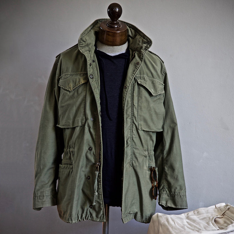 Olive green field jacket on a mannequin with a dark hoodie underneath, set in a softly lit room.
