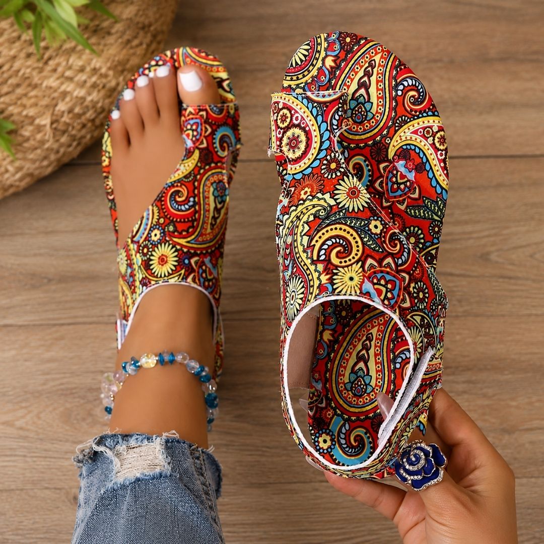 Colorful paisley-patterned orthopedic sandals on feet with anklet, denim jeans, wooden floor background.