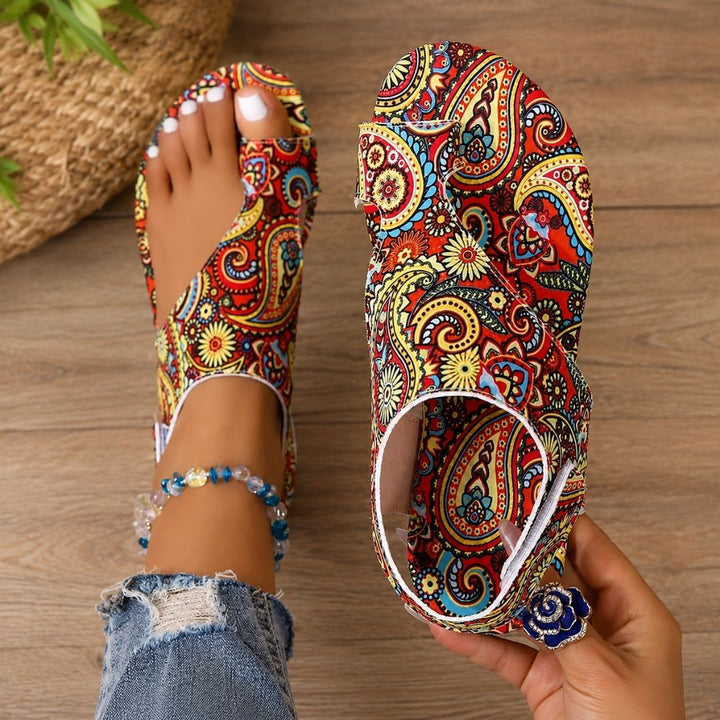 Colorful paisley-patterned orthopedic sandals on feet with anklet, denim jeans, wooden floor background.