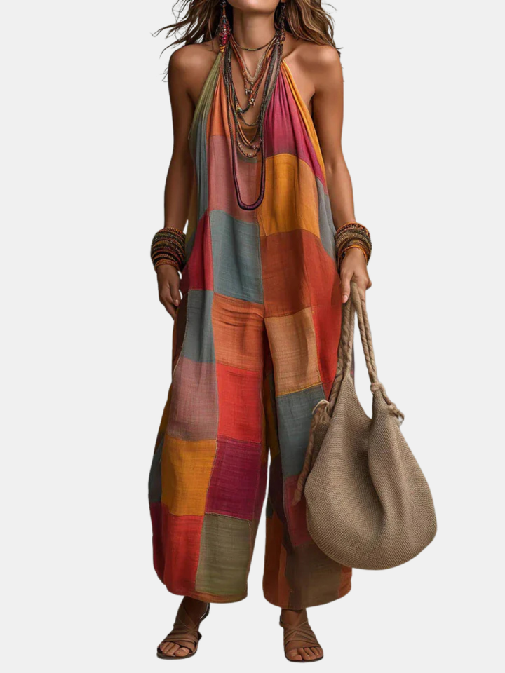 Bohemian jumpsuit in colorful patchwork, worn with layered necklaces and bangles. Model carries a woven bag, isolated on a white background.