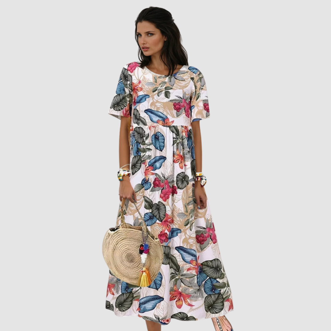 Woman wearing a flowing, floral summer dress in white with colorful flowers, holding a round straw bag, in a studio setting.