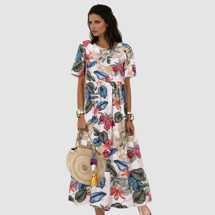 Woman wearing a flowing, floral summer dress in white with colorful flowers, holding a round straw bag, in a studio setting.