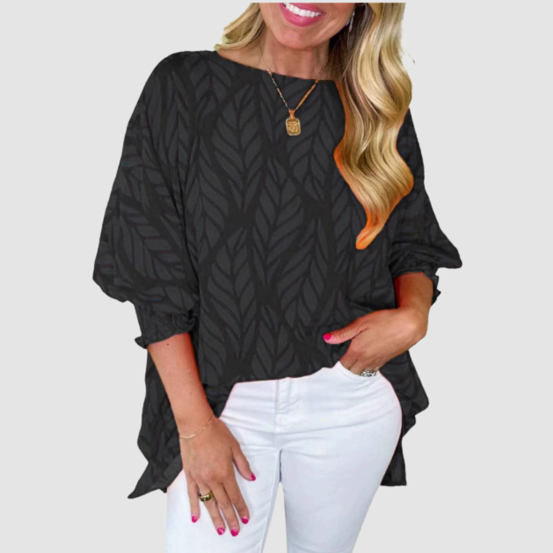 Woman wearing a black loose blouse with leaf pattern and white pants in a studio setting.