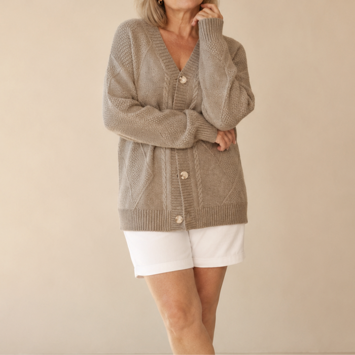 Person wearing a textured, brown knitted cardigan with buttons, paired with white shorts, in a minimalistic beige studio setting.