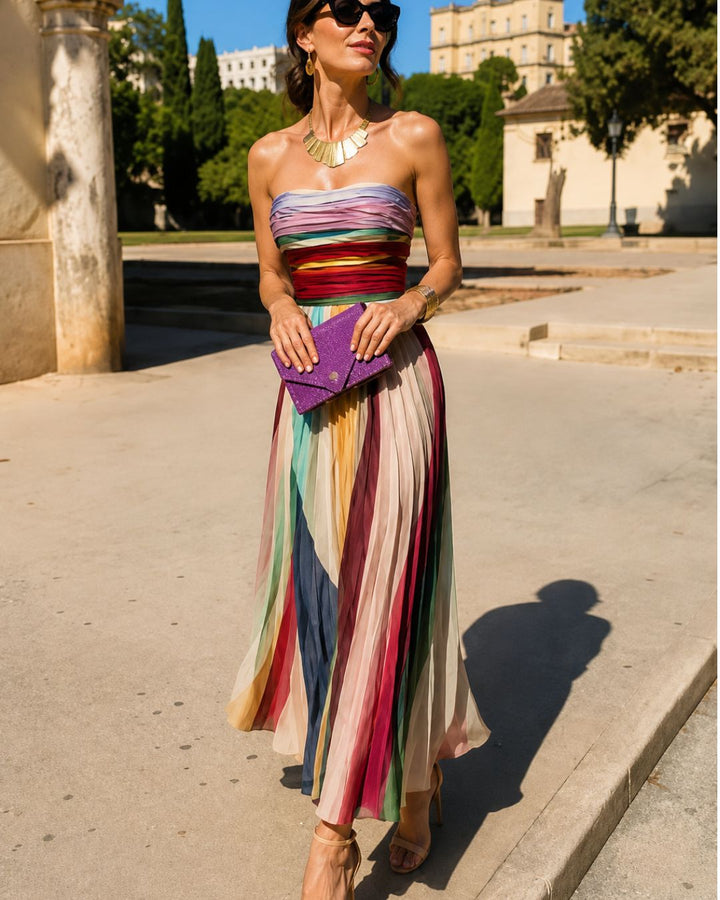 Woman wearing a colorful, strapless maxi dress with pleated details, holding a purple clutch, outdoors on a sunny day.