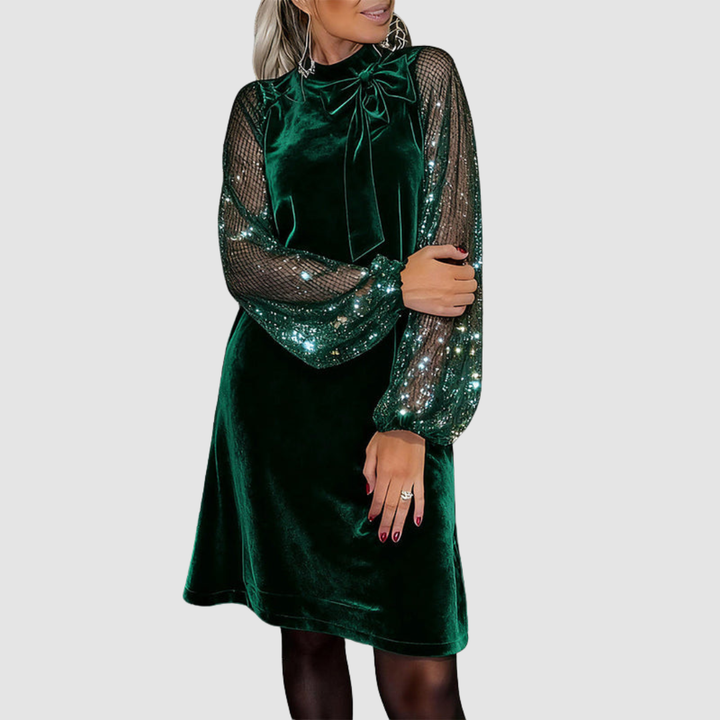 Green elegant dress with shimmering mesh sleeves, bow detail, and sequins, worn by a model, on a gray background.