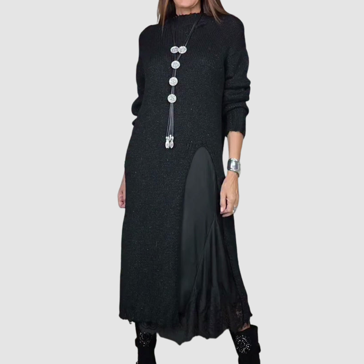 Woman wearing a long black dress with layered fabric and decorative necklace, posing against a plain gray background.