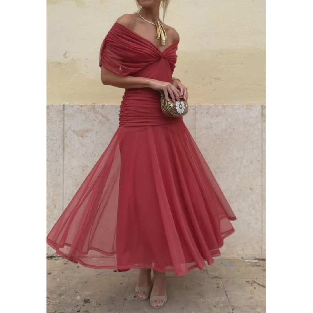 Woman in an elegant red mesh dress, holding a round clutch, posing against a light wall background.