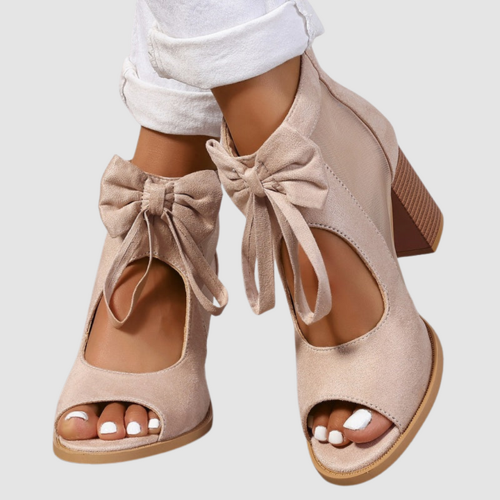Beige heeled sandals with bow detail, worn with cuffed white pants on a neutral background.