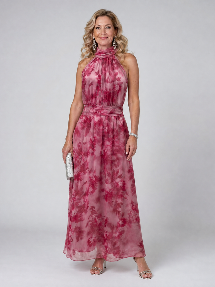 Woman in pink floral halter neck midi dress holding silver clutch, standing in studio setting.