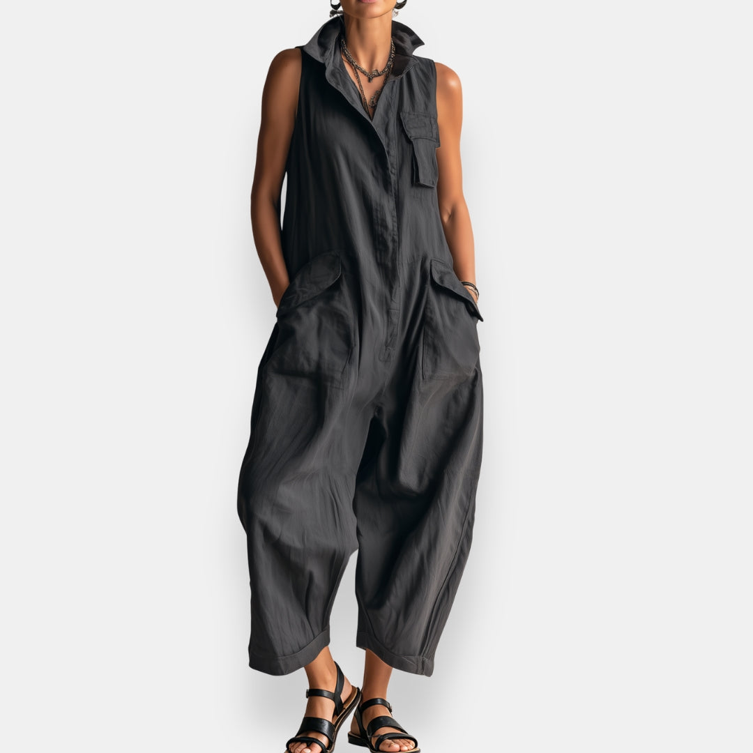 Black sleeveless cargo jumpsuit with collar and flap pockets, worn with strappy sandals in a studio setting.