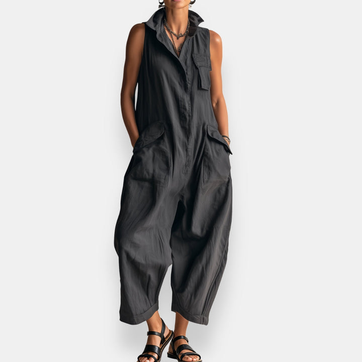 Black sleeveless cargo jumpsuit with collar and flap pockets, worn with strappy sandals in a studio setting.