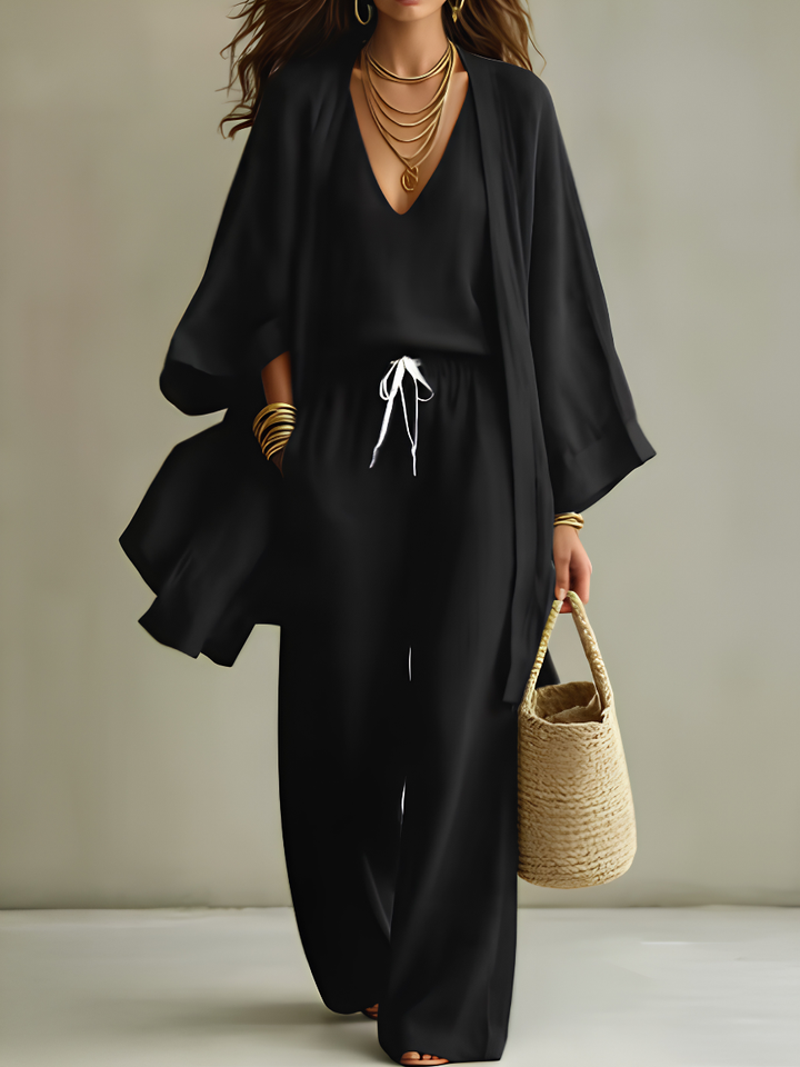 Black three-piece relaxed homewear set with v-neck top, drawstring pants, flowy overcoat, and woven bag in studio setting.