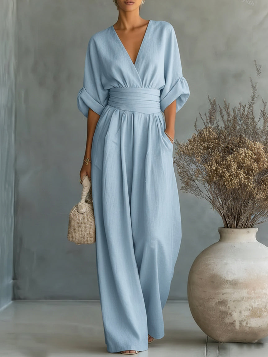 Light blue V-neck high-waist jumpsuit with rolled-up sleeves, modeled in a studio with neutral backdrop and large vase decoration.