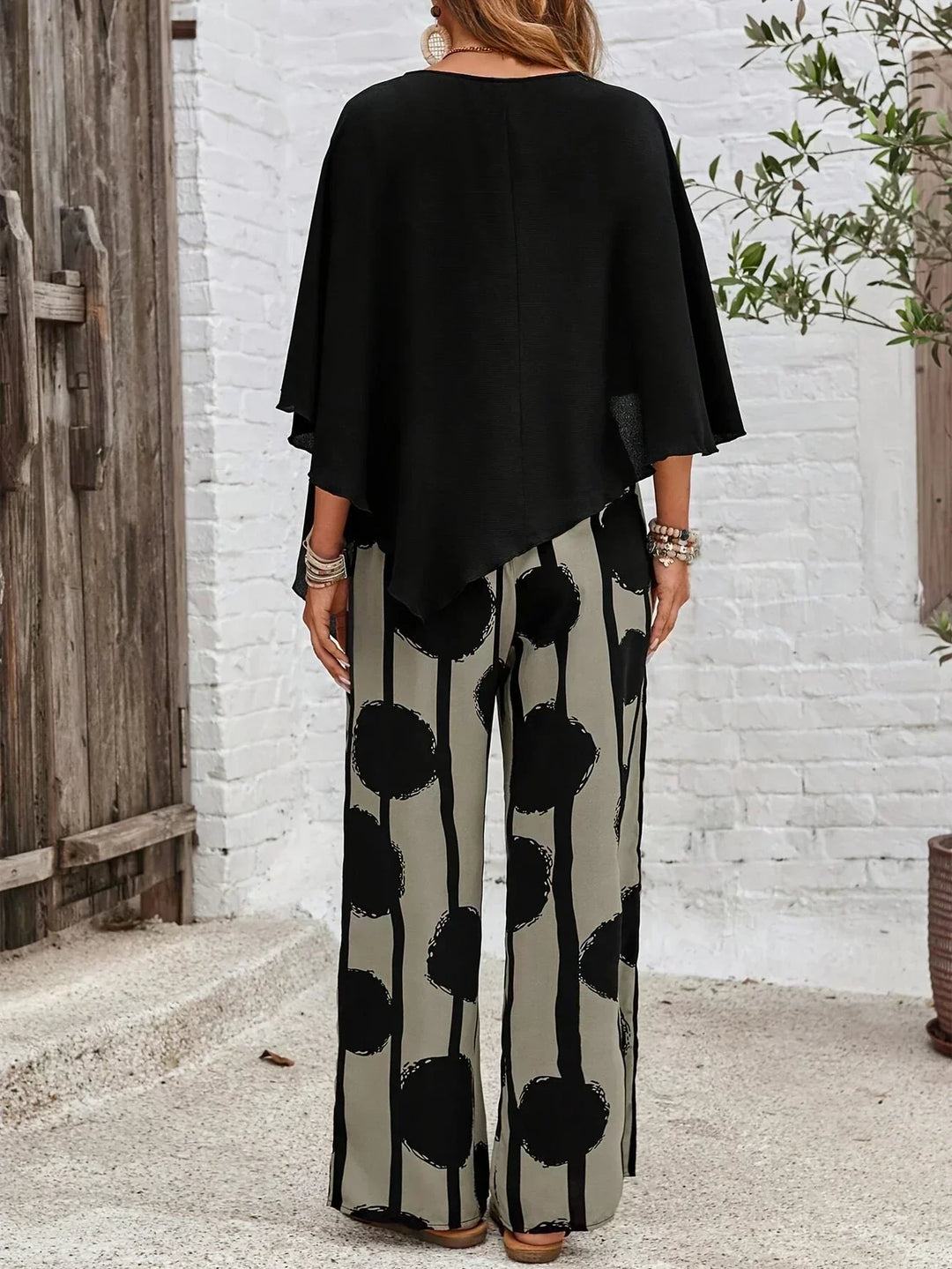 Back view of a black top and patterned wide-leg pants against a rustic outdoor backdrop with a white brick wall.