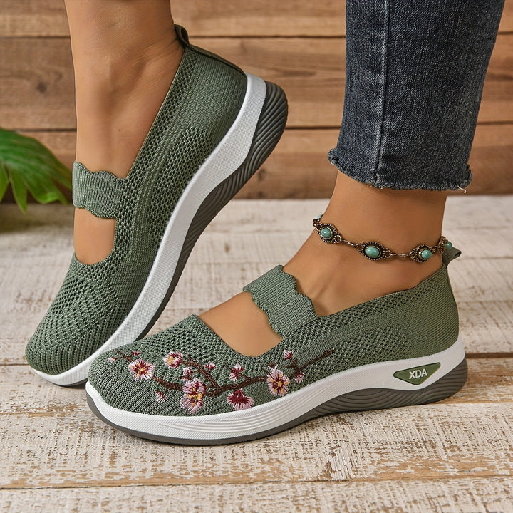 Side view of green orthopedic shoes with floral design, worn with jeans and anklet, on a wooden floor.