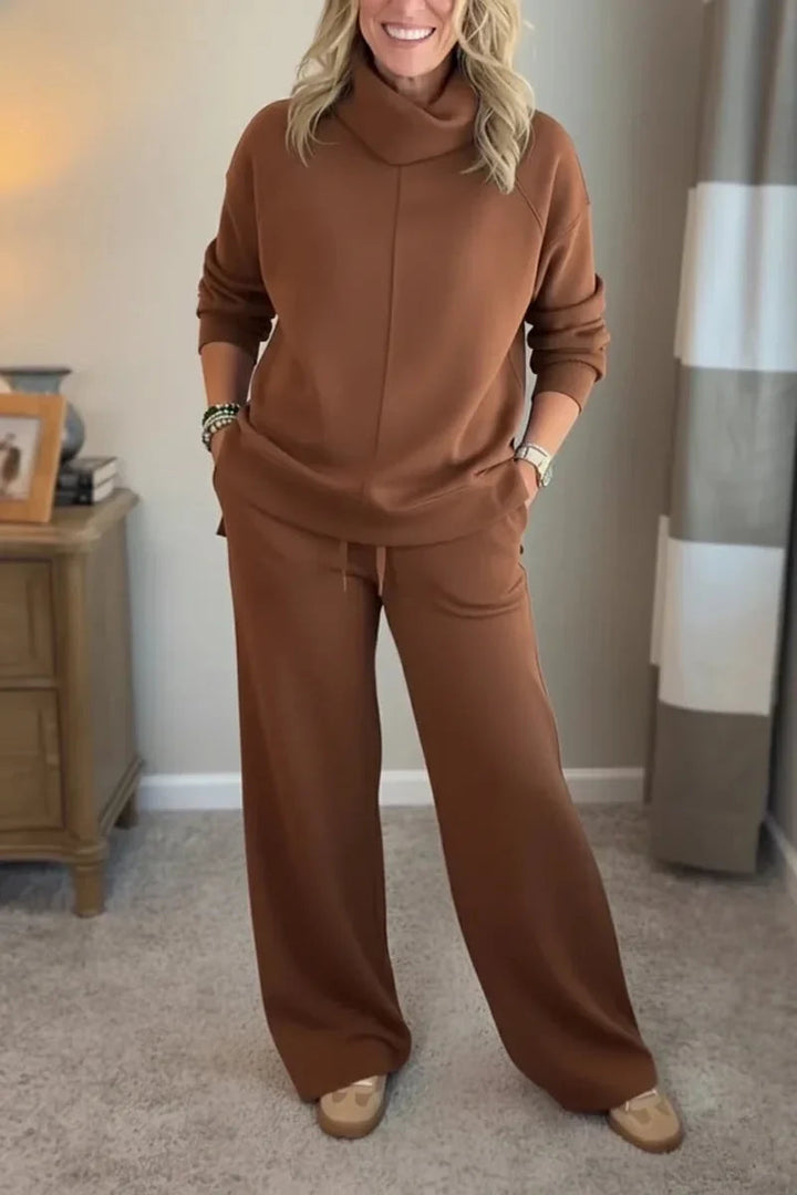 Woman in a brown comfortable set with long sleeves and wide-leg pants, standing in a carpeted room with a wooden table nearby.
