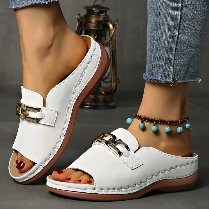 Stylish white sandals with gold chain details, worn indoors with jeans and anklet on grey flooring.