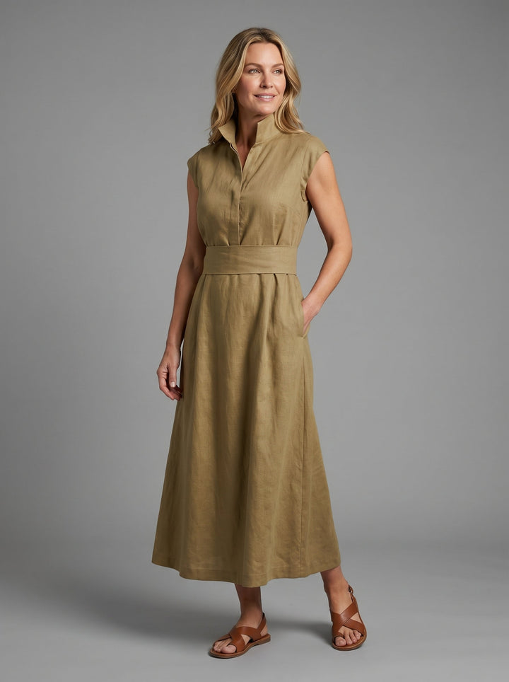 Smiling woman in khaki sleeveless dress with wide waistband, wearing sandals, posing in a studio with gray background.