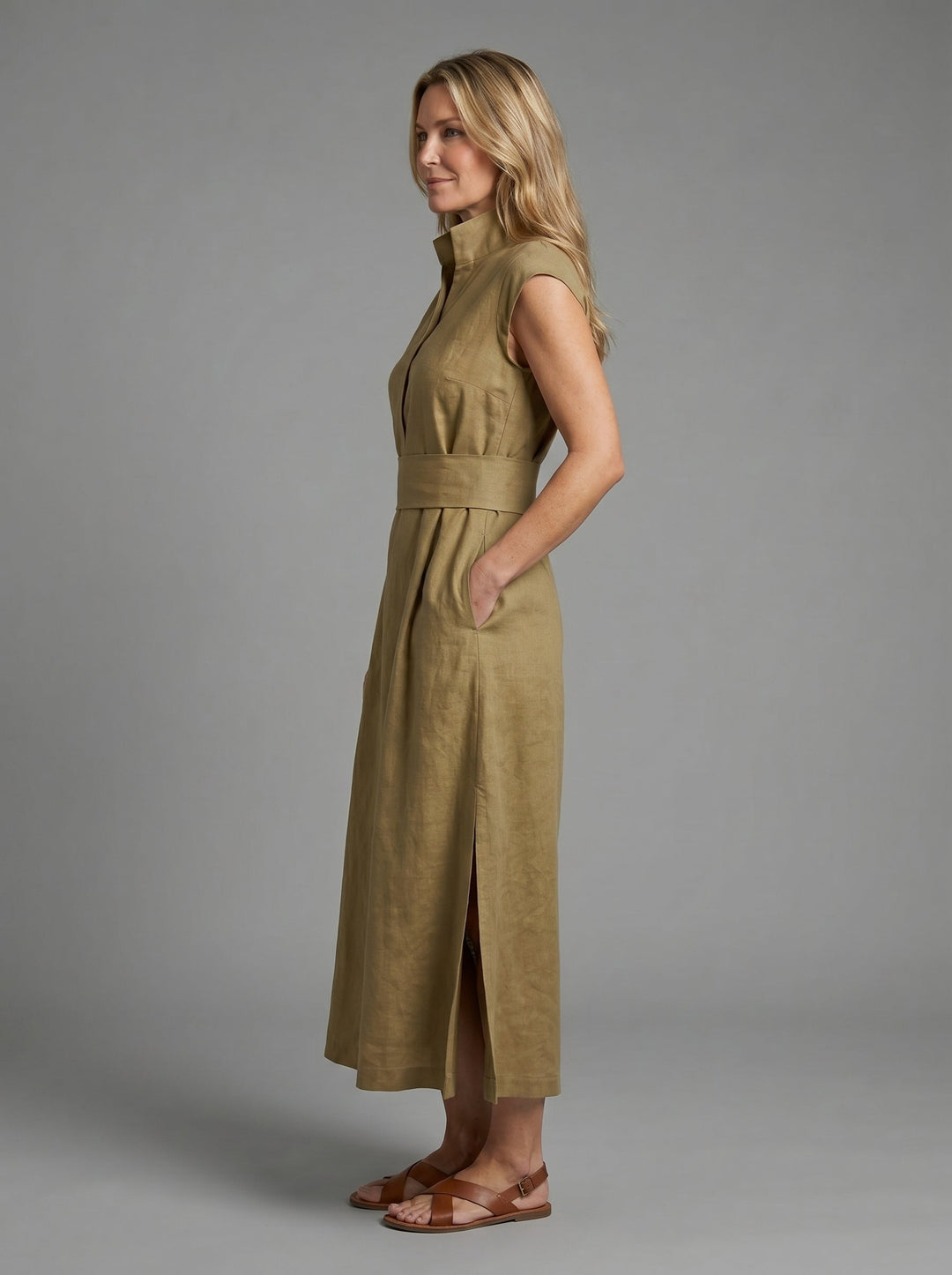 Side view of woman in khaki sleeveless dress with wide waistband, wearing sandals, posing in a studio with gray background.