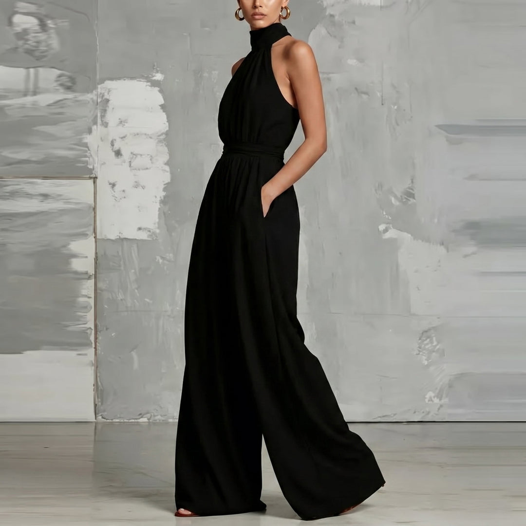 Model wearing a black halter-neck jumpsuit, hand in pocket, in a chic studio setting.