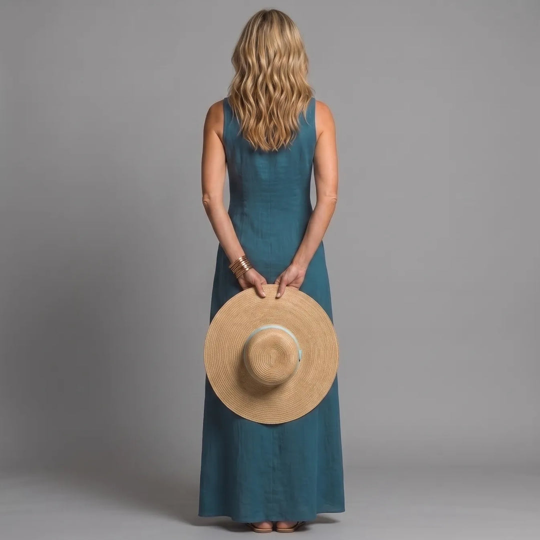 Back view of woman in teal maxi shirt dress, holding a straw hat, in a studio setting.