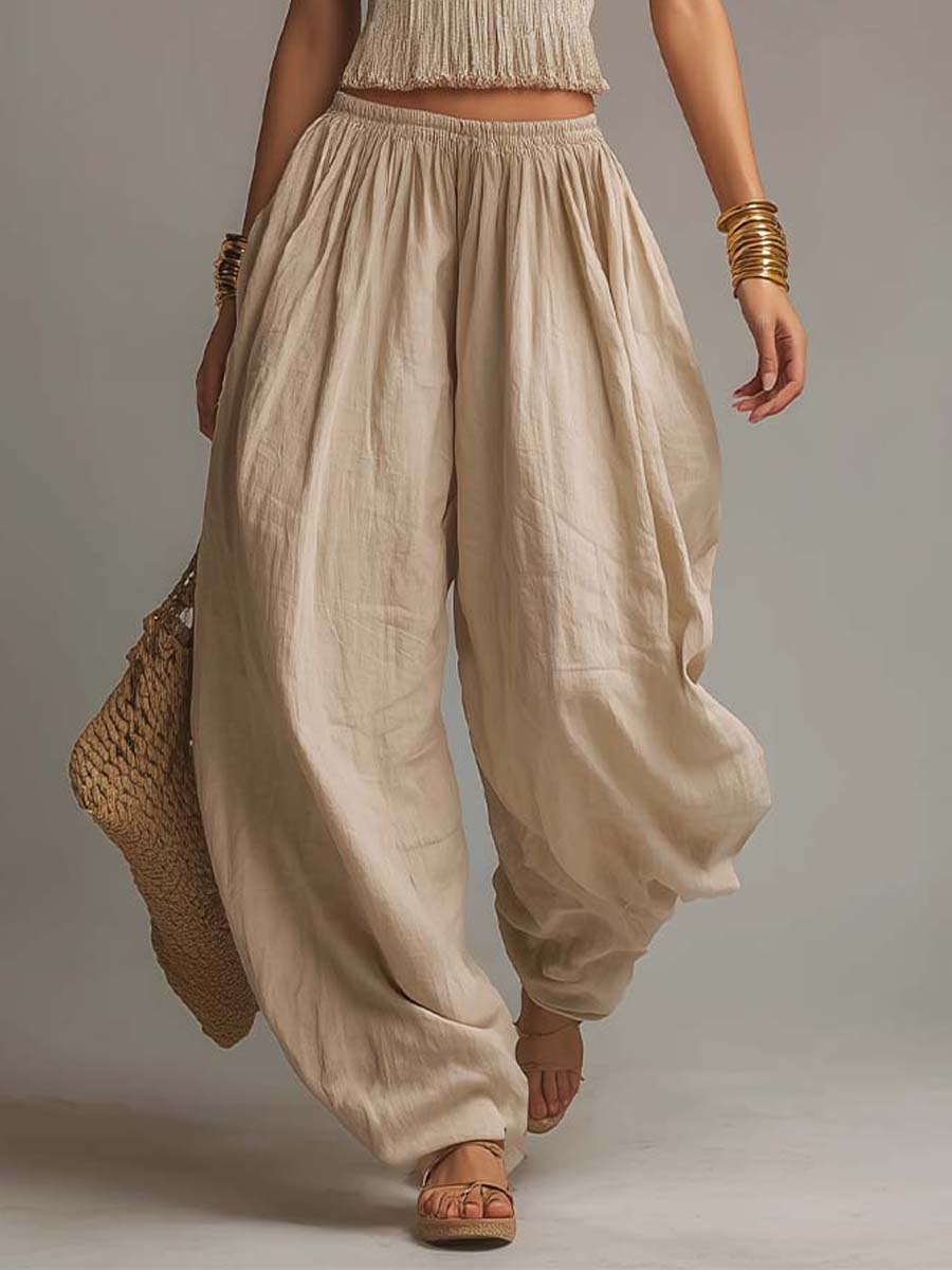 Close-up of beige flowing pants with a textured waistband, paired with sandals and gold bangles. Studio background.