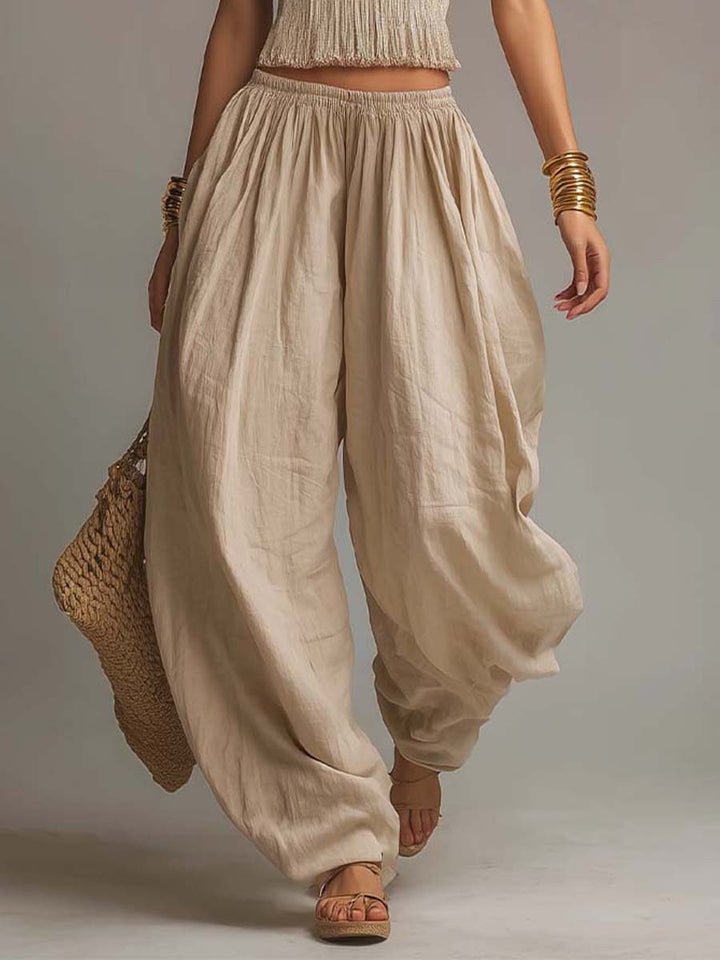 Close-up of beige flowing pants with a textured waistband, paired with sandals and gold bangles. Studio background.