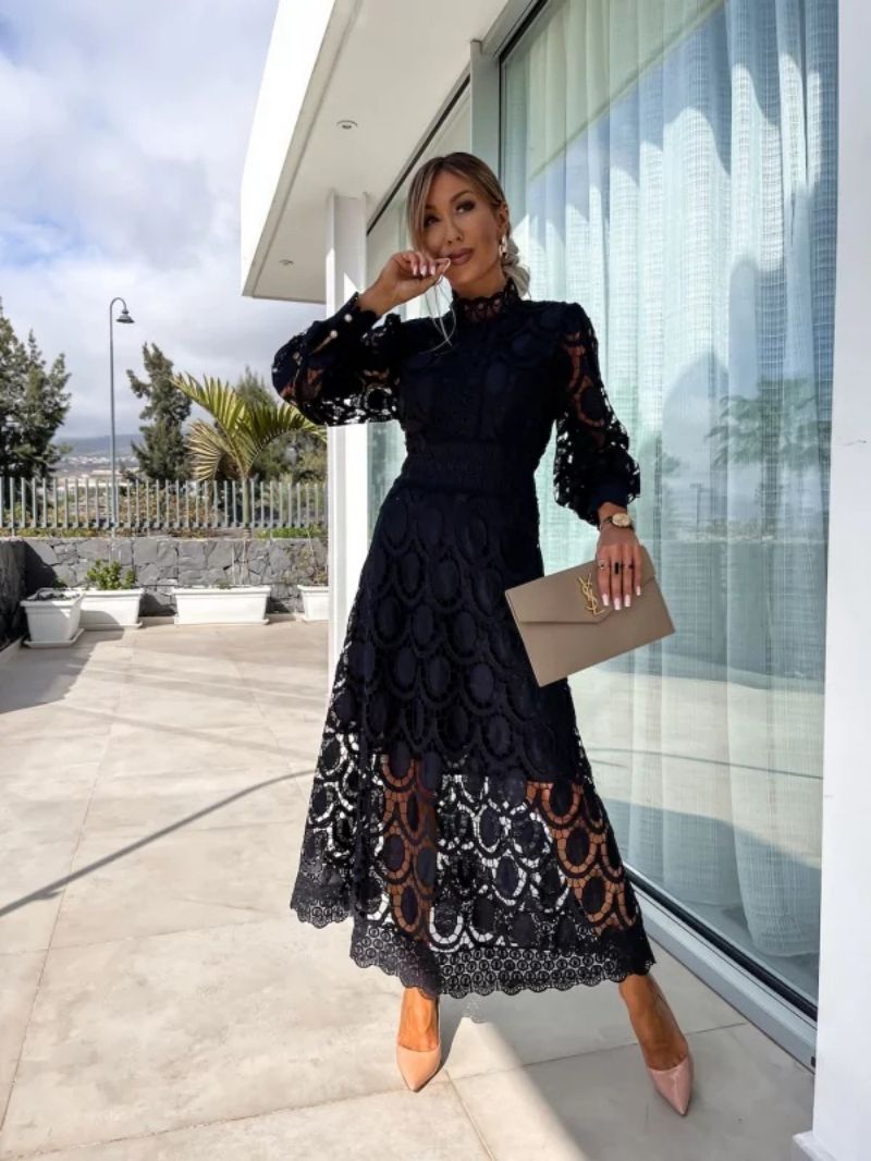 Black lace dress in an outdoor setting, accessorized with a beige clutch and nude heels, posing near a window with trees in the background.