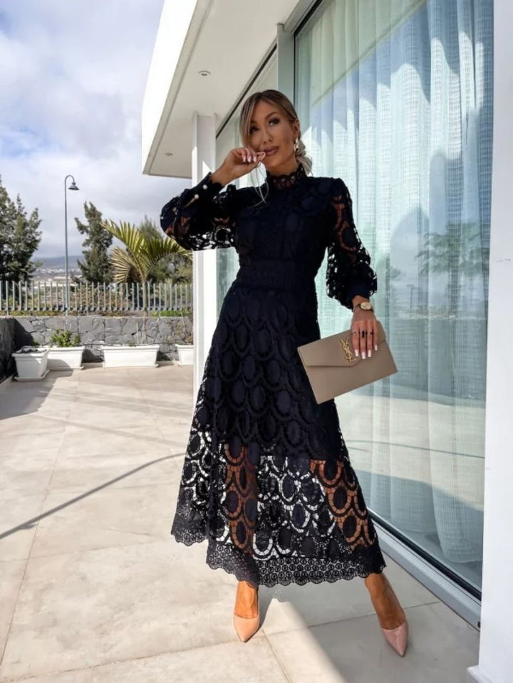 Black lace dress in an outdoor setting, accessorized with a beige clutch and nude heels, posing near a window with trees in the background.
