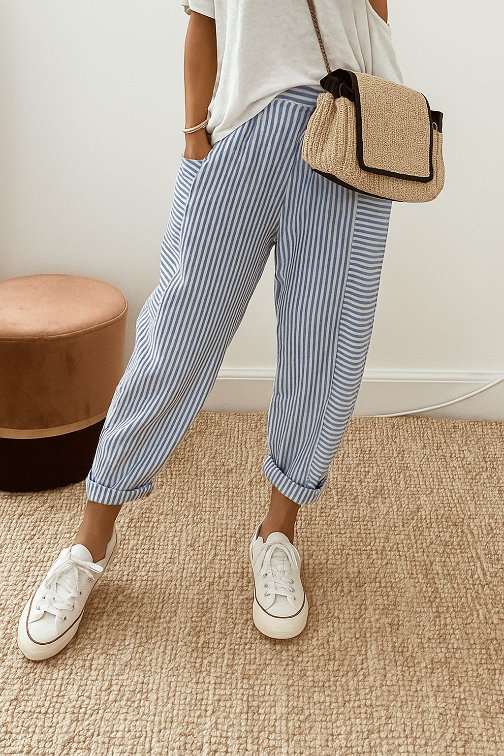 Person wearing blue striped pants with white sneakers and a casual white top, standing on a textured beige carpet near a brown stool.