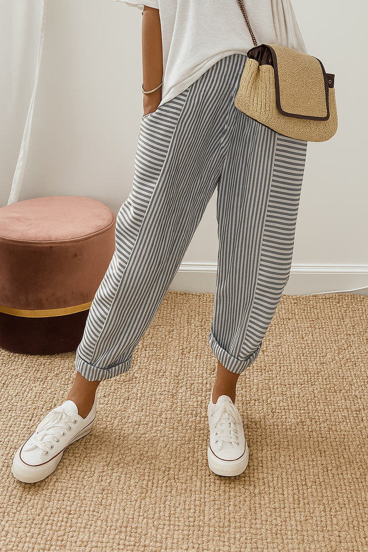 Person wearing gray striped pants with white sneakers and a casual white top, standing on a textured beige carpet near a brown stool.